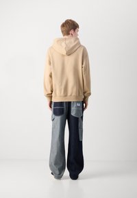 Hoodie in cotone beige con cucitura sul retro e cappuccio regolabile, abbinato a pantaloni in denim a gamba larga con toppe in toni di blu chiaro e scuro.