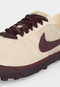 Παπούτσι Nike σε κρεμ απόχρωση με σκούρα καφέ κορδόνια, λογότυπο Swoosh σε σκούρο καφέ και σόλα σε σκούρο καφέ, ορατά σε λευκό φόντο.