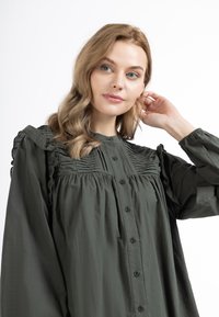 Groene, knoopsluiting blouse met geplooide details op de schouders, gestructureerde stof en gerimpelde accenten langs de mouwen. Beschikt over lange mouwen.