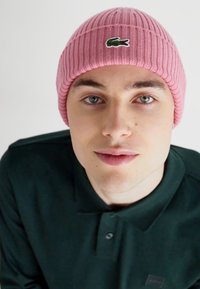 Ružový priliehavý čiapka s malým zeleným logom Lacoste. Model má na sebe tmavozelené polo tričko, ktoré zobrazuje textúru a strih čiapky.