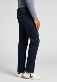 Mörkblå denimjeans med rak benlösning, med bakfickor och en svart läderetikett vid midjan, tillsammans med vita sneakers.