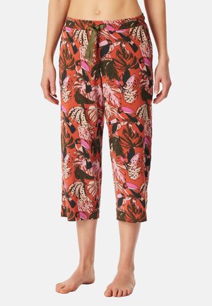 Frau trägt bauchfreie Loungebuxe mit pinkem, braunem und grünem tropischem Blatt- und Tukanspielfleck, grüner Kordel am Bund, barfuß.