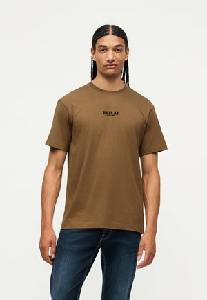 Jeune homme aux cheveux tressés portant un t-shirt marron à manches courtes avec le texte « REPLAY » et un jean bleu, debout devant un fond uni.