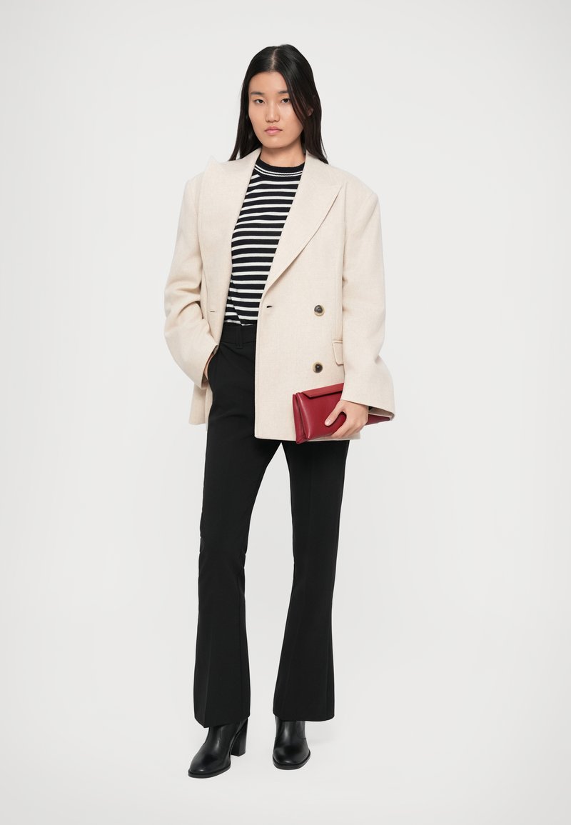 Blazer beige doppiopetto, top a righe black e white, pantaloni neri a zampa d'elefante e stivali neri con tacco. Tiene una borsetta rossa.