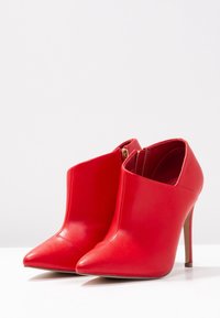 Botines de cuero rojo con punta puntiaguda, tacón de aguja alto y un diseño único con recortes, que incluyen una cremallera lateral para el cierre.