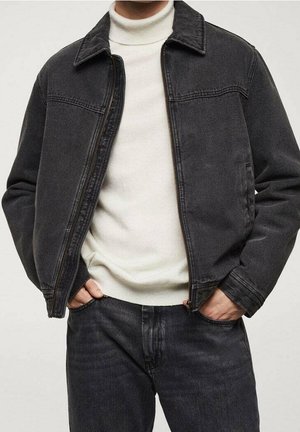 Mand iført en sort denimjakke over en hvid rullekravetrøje med hænderne i lommerne på matchende sorte denimjeans.