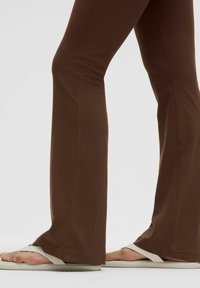 Braune, ausgestellte Hose aus dehnbarem Stoff mit auffälligen Nähten. Kombiniert mit elfenbeinfarbenen Flip-Flops, die ein einfaches Design zeigen.