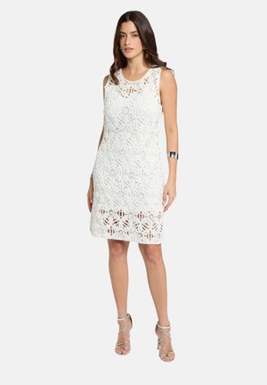 Donna che indossa un vestito bianco in pizzo senza maniche al ginocchio e sandali argentati con tacco alto, in piedi davanti a uno sfondo semplice.