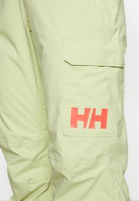 Svetlezelené cargo kalhoty mají velkou boční kapsu a výrazné červené logo "H/H". Textura vypadá hladce s matným povrchem.