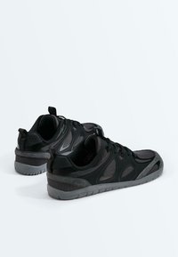 Paire de baskets athlétiques noires et gris foncé avec des superpositions en mesh et synthétiques, positionnées côte à côte sur un fond blanc.
