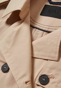 Col de trench beige avec des boutons noirs et des surpiqûres détaillées autour du col et des boutonnières, montrant une partie de l'étiquette à l'intérieur.