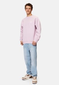 Lila huppari, jossa on pyöreä kaula-aukko ja ribbauskankaiset hihansuut, yhdistettynä vaaleansinisiin suoriin farkkuihin ja vaaleanruskeisiin lenkkareihin, joissa on ruskeita yksityiskohtia.