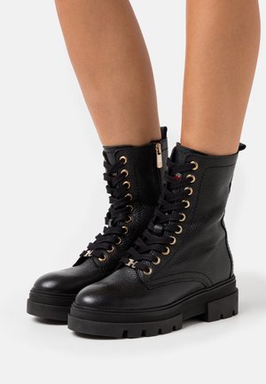 Stivaletti neri in pelle testurizzata con lacci, occhielli dorati, zip laterale, suola spessa e un piccolo dettaglio metallico a forma di "H" sui lacci, indossati sulle gambe.