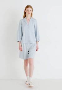 Camicia in lino azzurro chiaro con chiusura a bottoni e maniche a tre quarti, abbinata a pantaloni corti azzurro chiaro. Modello che indossa sneakers bianche.