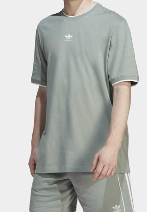 Mies pukeutunut vaaleanvihreään Adidas-paitaan ja siihen sopiviin shortseihin, joissa on valkoinen logo ja raidoituksia yksivärisellä taustalla.