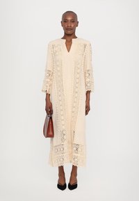 Beige long dress with intricate lace detailing, V-neckline, three-quarter sleeves, and a loose fit, paired with a brown handbag and black shoes.