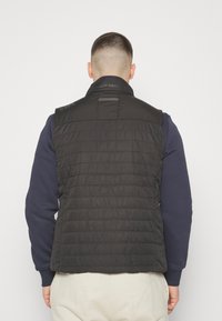 Černá péřová vesta s prošívaným designem, vysokým límcem a logem na zadní straně. Nositá přes námořnickou mikinu, ukázána zezadu.