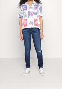 Camicia a maniche corte multicolore con stampe floreali e grafiche, abbinata a jeans blu slim fit con una leggera rottura e sneaker bianche.