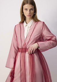 Robe rose transparente avec des accents rouges et une ceinture à nouer ; présente des manches longues et un design superposé et fluide sur une chemise blanche.