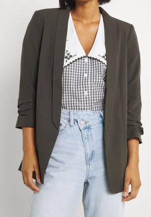 Vrouw draagt een olijfgroene blazer over een zwart-wit geruit blouse met een met bloemen geborduurde kraag, gecombineerd met lichtblauwe spijkerbroeken.