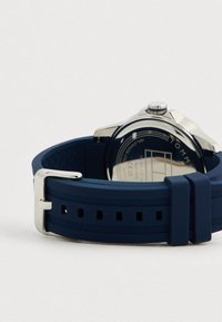 Hõbedane kellakorpus tumeinate sinise silikoonist rihmaga, metallist pandlaga ning graveeritud tagaküljel, kus on nähtavad tekst ja kaubamärgi andmed.