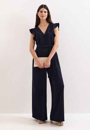Vrouw met lang donker haar die een mouwloze marineblauwe jumpsuit met ruches op de schouders draagt, houdt een beige clutch vast en draagt beige hakken.