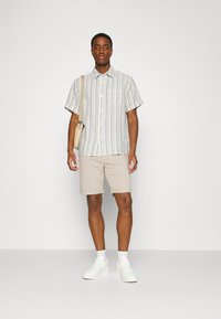 Button-up short-sleeve shirt with blue and cream stripes, beige shorts, and white sneakers. Model holding a light-colored shoulder bag.