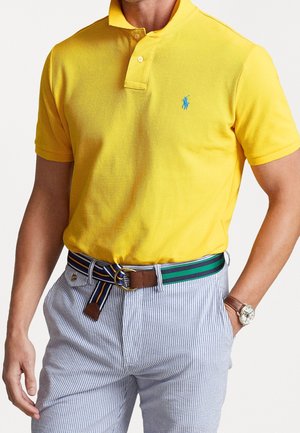 Mann trägt gelbes Poloshirt mit blauem Logo, blau-weiß gestreifte Hose, grün-blau gestreiften Gürtel und Armbanduhr, steht mit einer Hand in der Tasche.