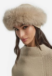 Fuskpälsmössa i beige med ett brett fluffigt band. Stickat material på undersidan med en mjuk textur och rundad form.