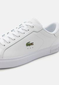 Sneaker bianca realizzata in pelle liscia, caratterizzata da un logo di coccodrillo ricamato in verde, punta rotonda e lacci bianchi piatti su una suola in gomma bianca.