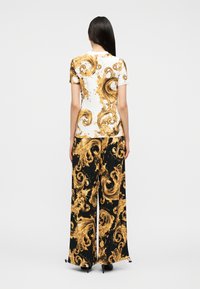 Γυναίκα με μακριά σκούρα μαλλιά, ντυμένη με λευκή και μαύρη ένδυση με περίτεχνα χρυσά μπαρόκ στροβιλισμένα σχέδια, στέκεται μπροστά σε απλό ανοιχτό φόντο.