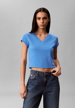 Kobieta ubrana w dopasowany niebieski krótki top z dekoltem w serek oraz luźne granatowe jeansy z wysokim stanem na jednolitym szarym tle.