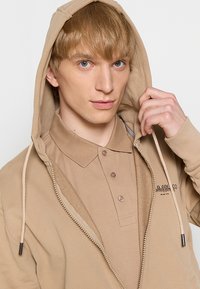 Beige zip-up hette med snøring, med en teksturert innvendig og en krage fra en matchende beige poloskjorte under.