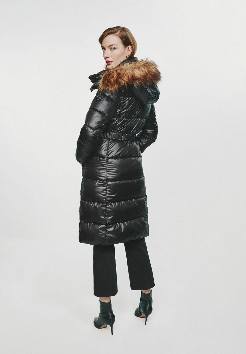 Kobieta w długim czarnym płaszczu puchowym z kapturem wykończonym futrem, w czarnych spodniach 3/4 i czarnych botkach na wysokim obcasie, stojąca bokiem.