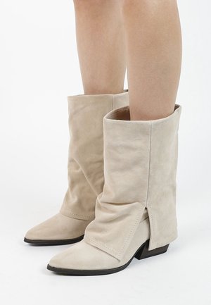 Bottes mi-mollet en daim beige avec bouts pointus, talons bas noirs, et un design plié légèrement affaissé, portées sur jambes nues.