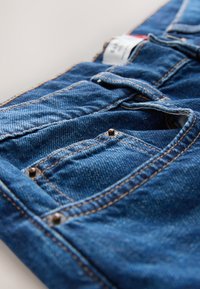 Nahaufnahme von gefalteten blauen Denim-Jeans, die die Vordertasche, das kleine Münzfach, Nähte und Metallnieten vor hellem Hintergrund zeigen.