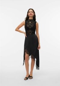 Melna kleita ar mežģīņu bodiju, bez piedurknēm un asimetrisku maliņu ar mežģīņu apdari. Gluda auduma, potītes garuma, valkāta ar atvērtām kurpēm.