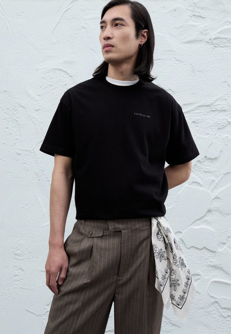 Svart T-skjorte med korte ermer med subtil tekst "Last Resort Age." Kombinert med brune bukser med nålestriper og et hvitt bandana med blomsterpattern.