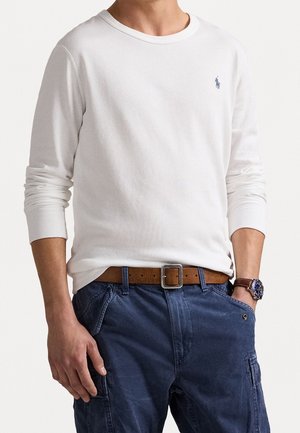 Mies, jolla on valkoinen pitkähihainen paita, jossa pieni logo, siniset cargo-housut, ruskea nahkavyö ja rannekello, seisoo käsi taskussa.