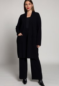 Cardigan noir ouvert à l'avant avec des manches longues et deux poches avant, fabriqué en matériau doux et texturé, assorti avec un pantalon noir à jambes larges.