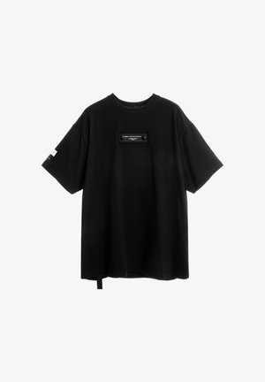 Camiseta negra oversize de manga corta con escote redondo, pequeño parche con texto en el pecho y gráfico blanco en la manga izquierda.