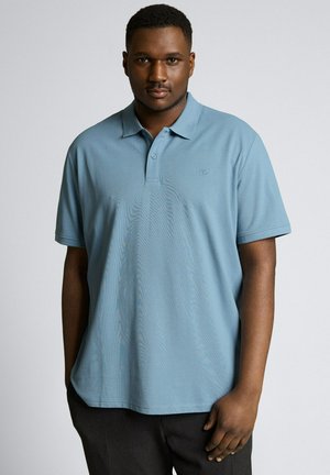 Mann mit kurzen Haaren, der ein hellblaues kurzärmeliges Polohemd und dunkle Hosen trägt, steht vor einem schlichten hellen Hintergrund.