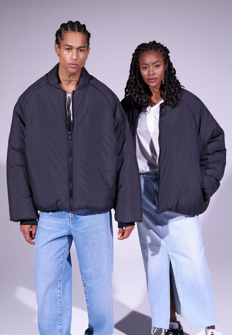Jaquetas bomber acolchoadas negras em dois modelos, com fecho de correr na frente, ajuste oversized e punhos canelados, combinadas com jeans de denim azul claro.