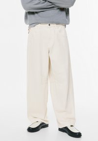 Videbenede, kreme-farvede bukser lavet af bomuld, med subtile syninger og klassiske jeanslommer, parret med sorte loafers.