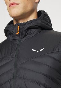 Veste isolée noire avec une capuche, présentant une texture matelassée verticale, une fermeture éclair avant et un logo blanc sur la poitrine. Accentuée par des détails en orange.