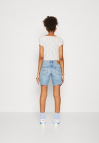 Valkoinen paita, jossa on pieniä mustia pisteitä, siniset farkkushortsit ja siniset sukat valkoisilla kengillä. Keskittymisenä takaosan näkymä, joka esittelee vaatteiden yksityiskohtia.