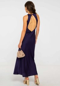 Mujer vestida con un vestido sin mangas de color azul marino y espalda descubierta, sosteniendo un bolso beige y llevando sandalias de tacón alto doradas, de espaldas.