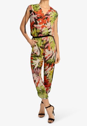 Kvinde iført ærmeløs buksedragt med blomsterprint i grønne, røde, rosa og hvide blade, sort bælte og sorte ankel-rem hæle, stående med hånden i lommen.