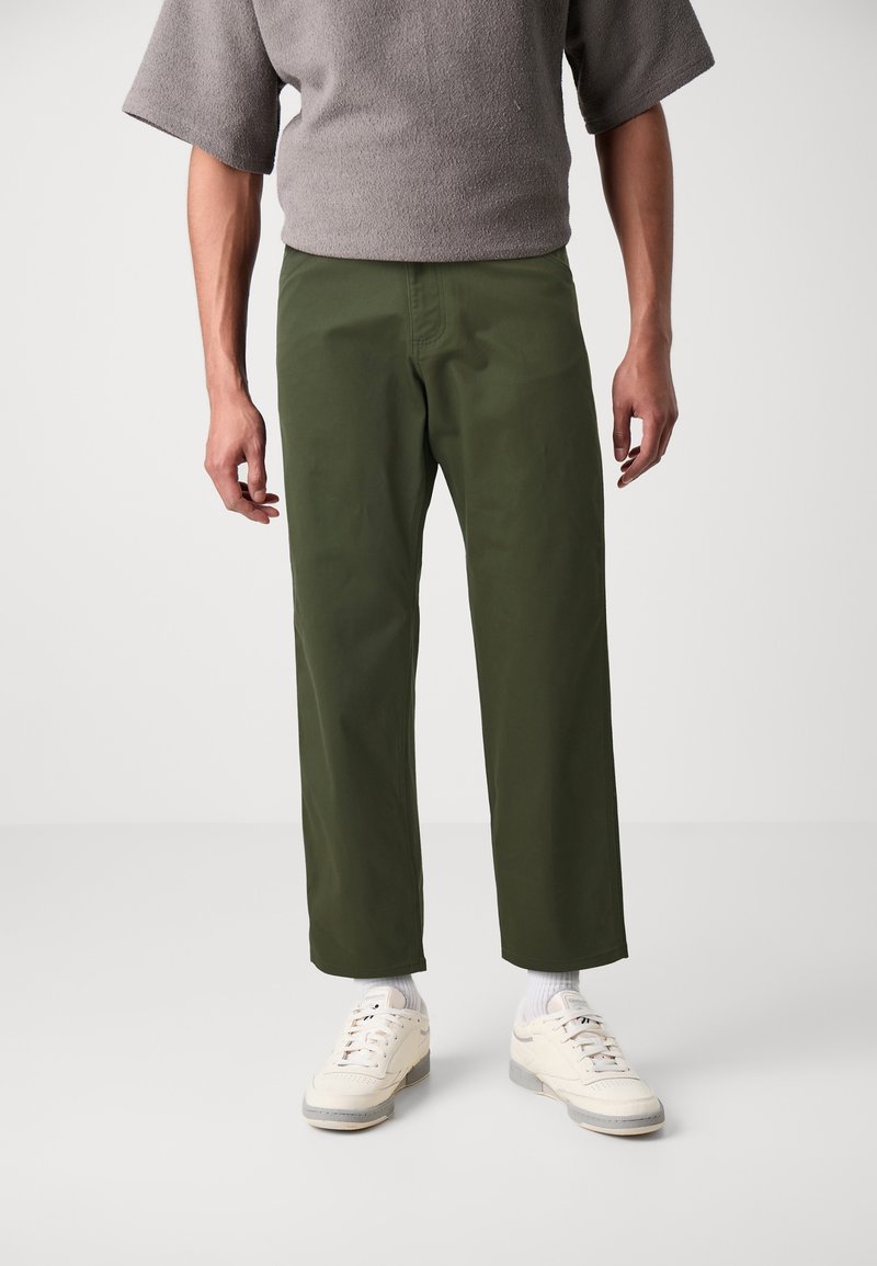 Personne portant un pantalon droit vert olive, des chaussettes blanches et des baskets beiges, debout devant un fond clair uni.