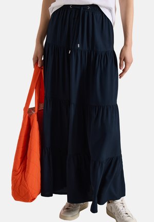 Długa granatowa spódnica na warstwach z sznurkiem w talii, noszona z białymi trampkami i trzymająca dużą pomarańczową pikowaną torbę.